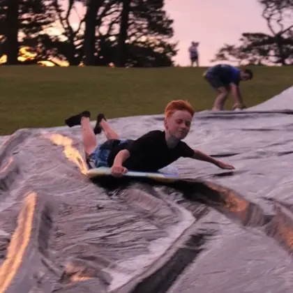 A teenager riding a bodyboard down a slip-and-slide on the grass at dusk