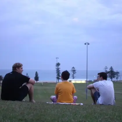 Three teenagers sitting on a grassy hill looking out to the ocean at dusk