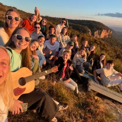 Smiling group selfie of teenagers on a grassy hillside at sunset, one holding a guitar