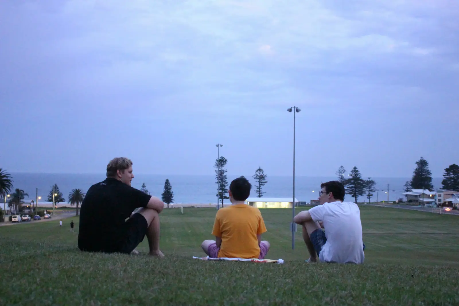 Three teenagers sitting on a grassy hill looking out to the ocean at dusk