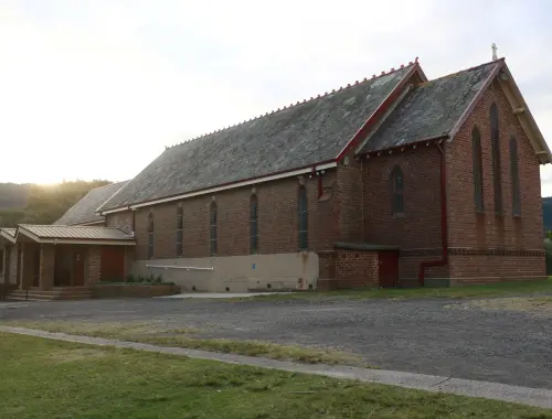 The red-brick exterior of Bulli Anglican Church, with its slate roof and arched windows, in late afternoon light