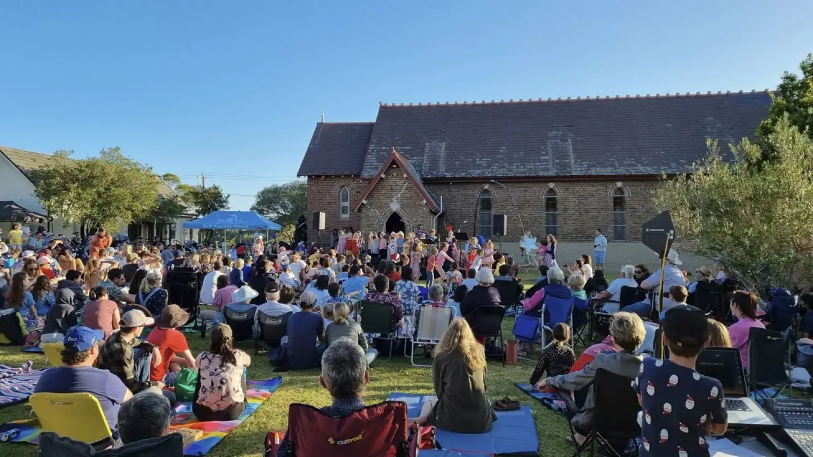 A large crowd on picnic rugs and camp chairs in front of the brick church, watching a children's choir perform at an outdoor carols event
